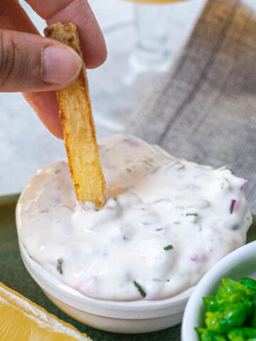 A hand dipping a crispy french fry into a small bowl of creamy white sauce garnished with herbs and diced onions, placed on a table with a linen napkin.