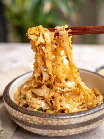 Chopsticks lifting spicy noodles with sesame seeds from a ceramic bowl, focusing on the texture and steam of the freshly cooked dish.
