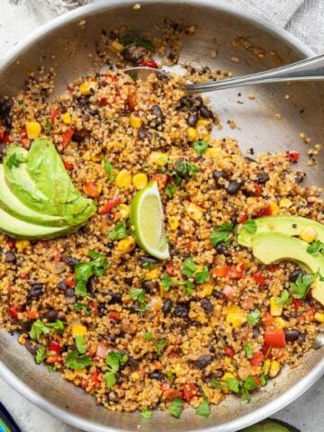 A colorful quinoa salad in a bowl with black beans, corn, diced tomatoes, chopped peppers, cilantro, lime wedge, and sliced avocado, served with a spoon.
