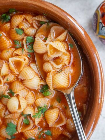 A close-up image of a traditional terracotta bowl filled with a hearty pasta stew, featuring conchiglie pasta in a rich, reddish sauce sprinkled with fresh parsley. a spoon is partially submerged in the dish.