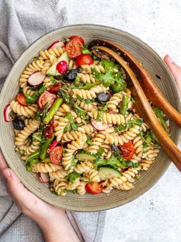 A person holds a bowl of colorful pasta salad with fusilli, cherry tomatoes, black olives, radishes, asparagus, and arugula. The salad is garnished with seeds. Wooden salad tongs rest in the bowl, and a light grey cloth is partially visible.