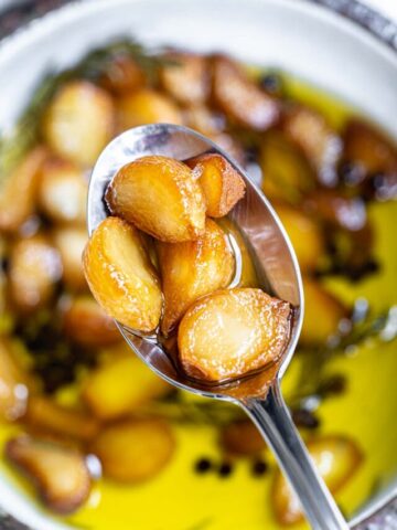 A close-up of a spoon holding several caramelized roasted garlic cloves above a bowl filled with more roasted garlic cloves soaking in olive oil. The background shows blurry garlic cloves and sprigs of herbs in the oil.