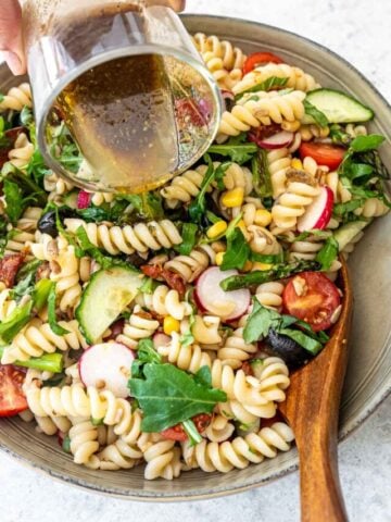 A bowl of pasta salad with colorful ingredients like spiral pasta, radishes, cucumbers, tomatoes, and leafy greens. A person is pouring a dressing from a glass container over the salad. A wooden spoon is in the bowl, ready for mixing.
