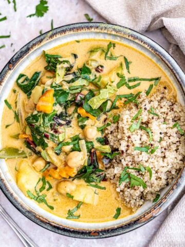 A bowl filled with vibrant vegetable curry made with chickpeas, leafy greens, and assorted vegetables in a creamy, yellow curry sauce. A serving of quinoa is placed beside the curry. The dish is garnished with chopped fresh herbs and set on a light-colored surface.