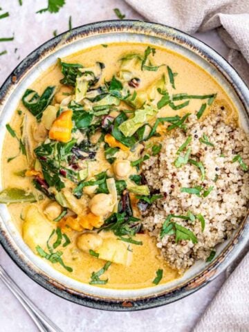 A bowl of vibrant, creamy vegetable curry featuring mixed greens, chickpeas, and colorful vegetables, garnished with fresh herbs. To the side, there is a portion of cooked quinoa, also sprinkled with herbs. A white cloth napkin and a part of a fork are visible nearby.